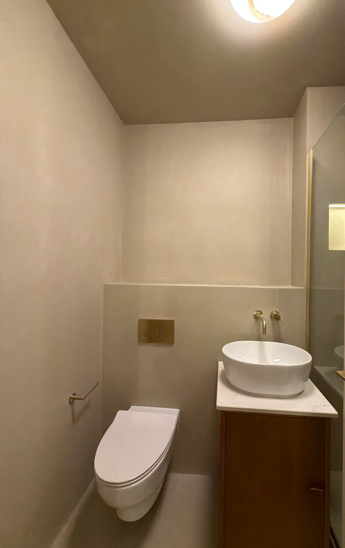 Narrow cloakroom with a wall-hung toilet, vessel basin and brass tap against pale warm-grey walls