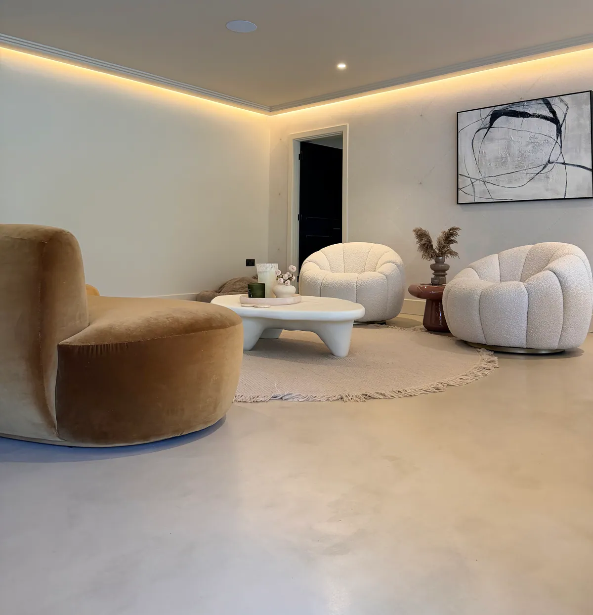Low view of a pale microcement floor in a living area with curved seating, a round rug and cove lighting