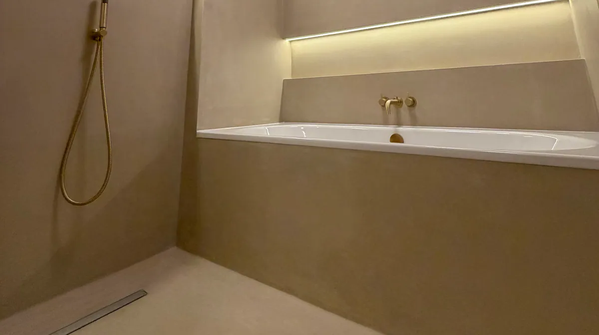 Bath beneath a lit wall niche, with a brass hand shower and a linear drain in the floor