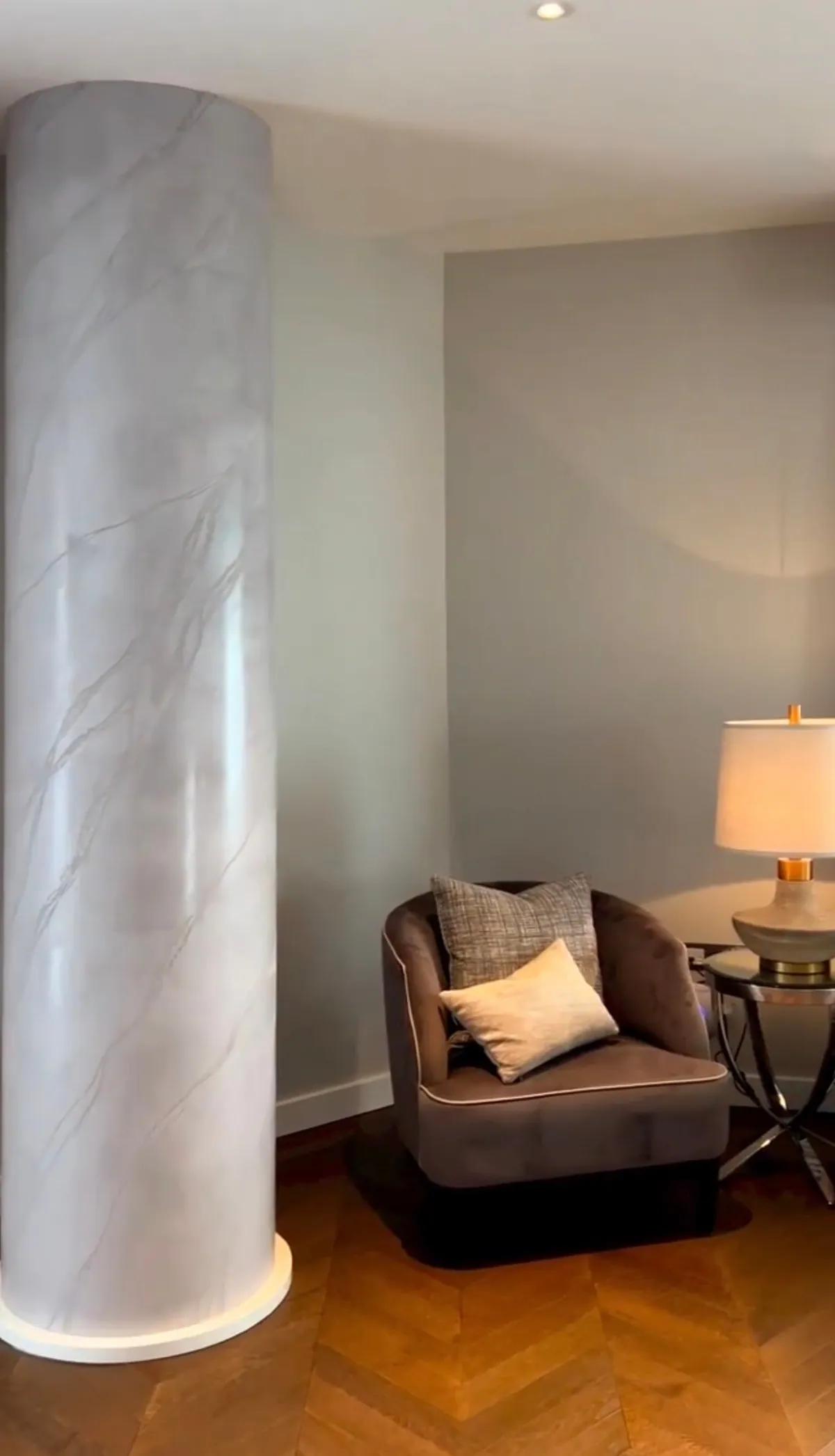 Pale veined column beside an armchair, a table lamp and a herringbone timber floor