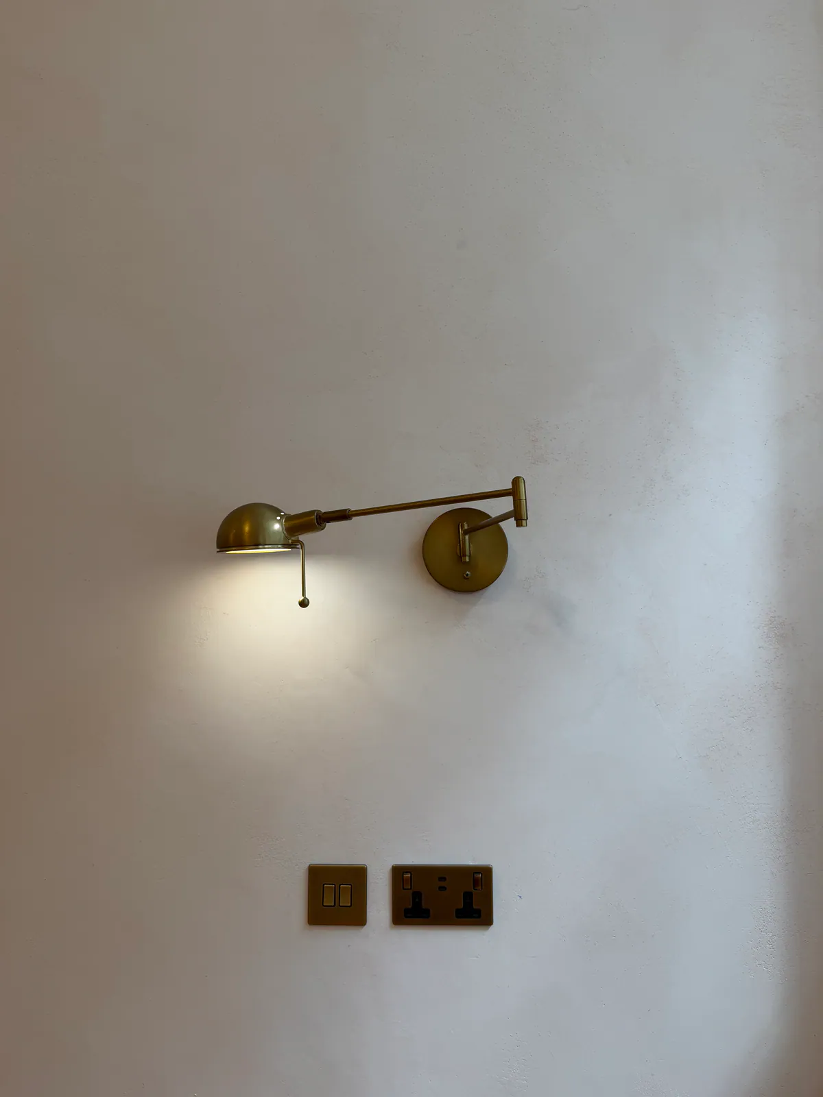 Brass swing-arm wall light above a brass switch and double socket on a pale plaster wall