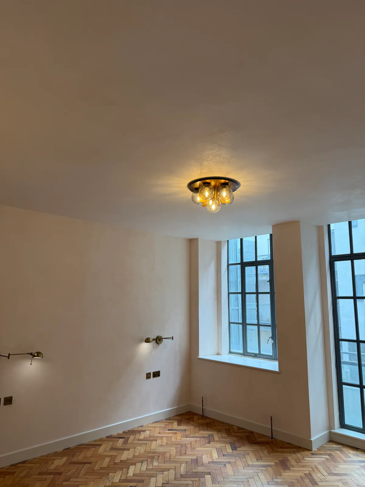 Empty room with plastered walls and ceiling, brass wall lights, steel-framed windows and a herringbone floor